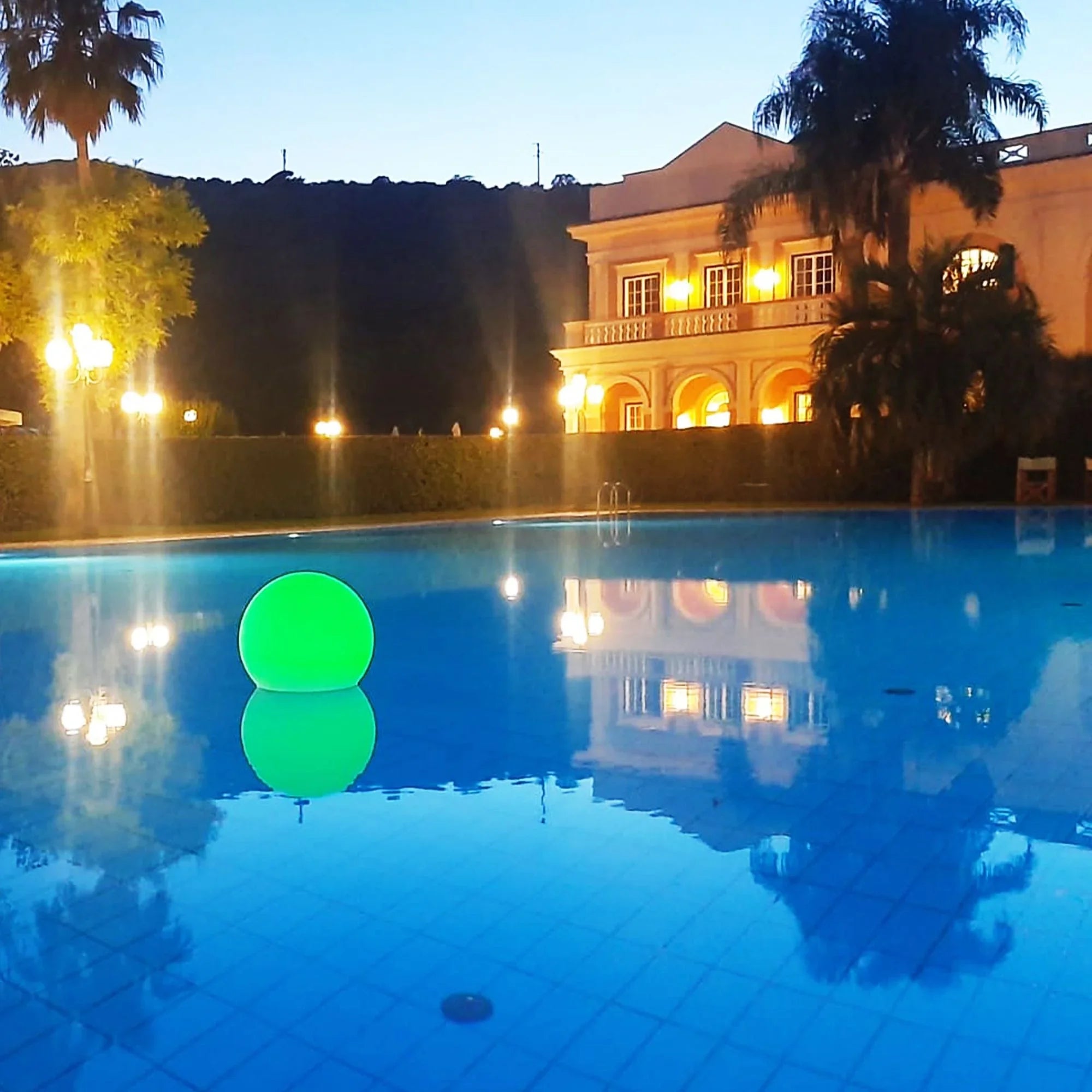 Piscina serale con palla luminosa verde, villa sullo sfondo e palme, atmosfera elegante